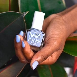 A close up of a hand cradling a bottle of So Fly nail polish from Hello Birdie which is a sky blue hue. The bottle is cube shaped with a white cap and text and the model is wearing the polish on the nails. The background has rubber plant leaves.
