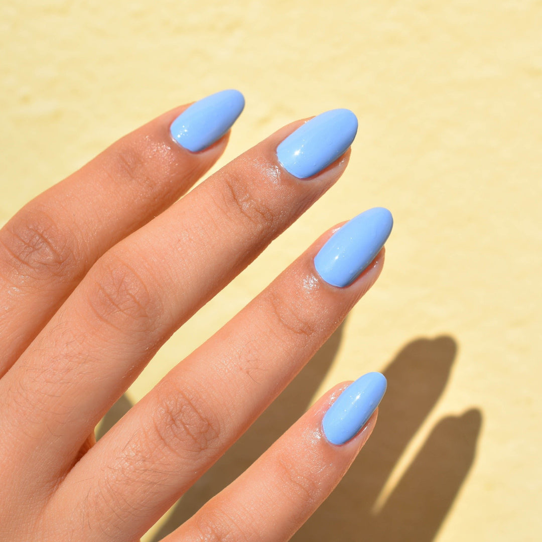 A close up of fingers wearing So Fly nail polish from Hello Birdie which is a bright sky blue. The medium toned fingers are illuminated with daylight against a soft focused yellow wall.