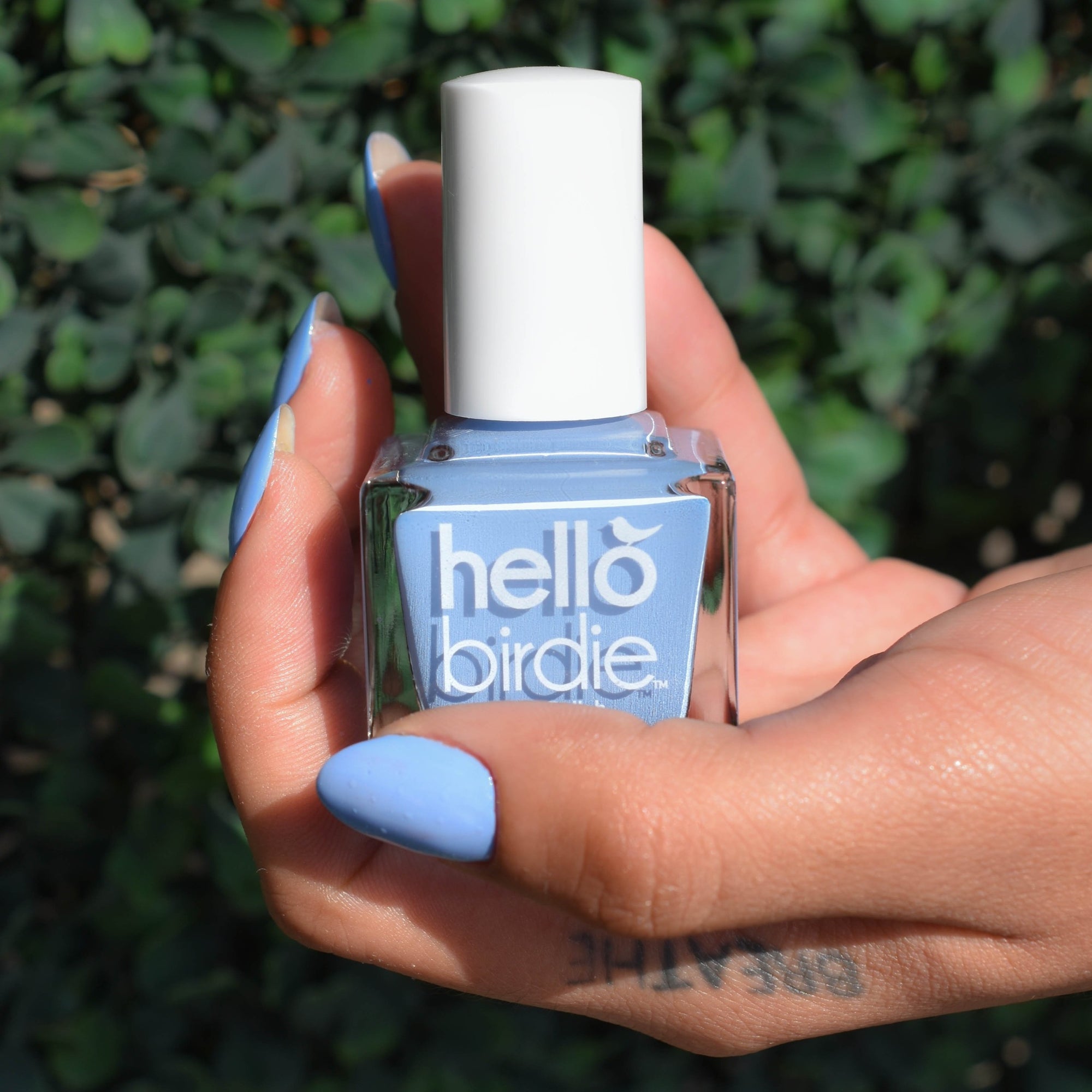 A close up of medium toned hand holding a bottle of So Fly nail polish from Hello Birdie which is a bright sky blue hue. The nails are painted with the polish and a tattoo saying "breathe" is revealed on the side of the hand. The background is an out of focus ivy wall.