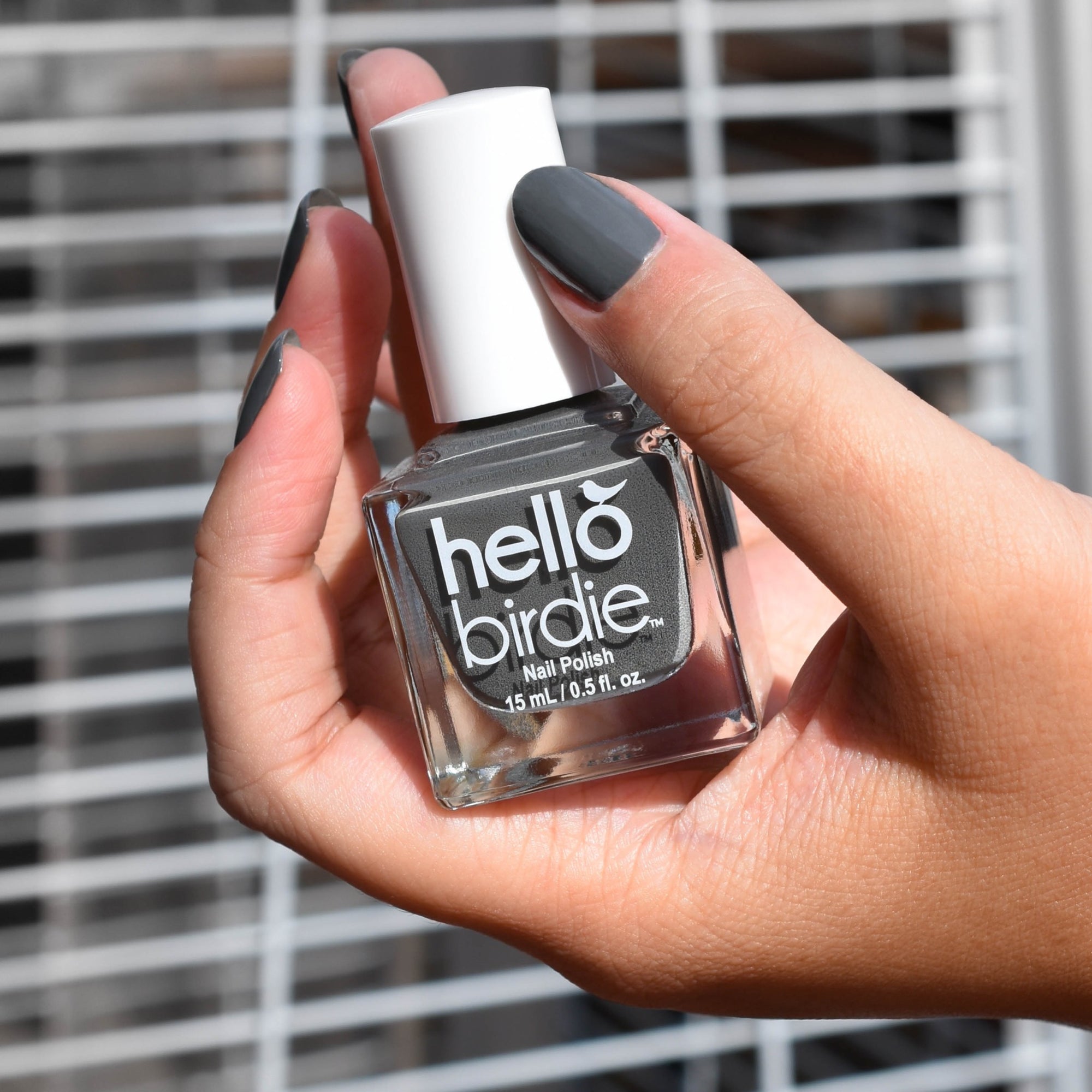A close up a midtoned hand comes in from the right side holding a bottle of Stormy Feather nail polish from Hello Birdie and wearing the same color on the nails. The polish hue is a dark gray and the bottle has white logo and white cap with cube shape. The background is out of focus silver cage.
