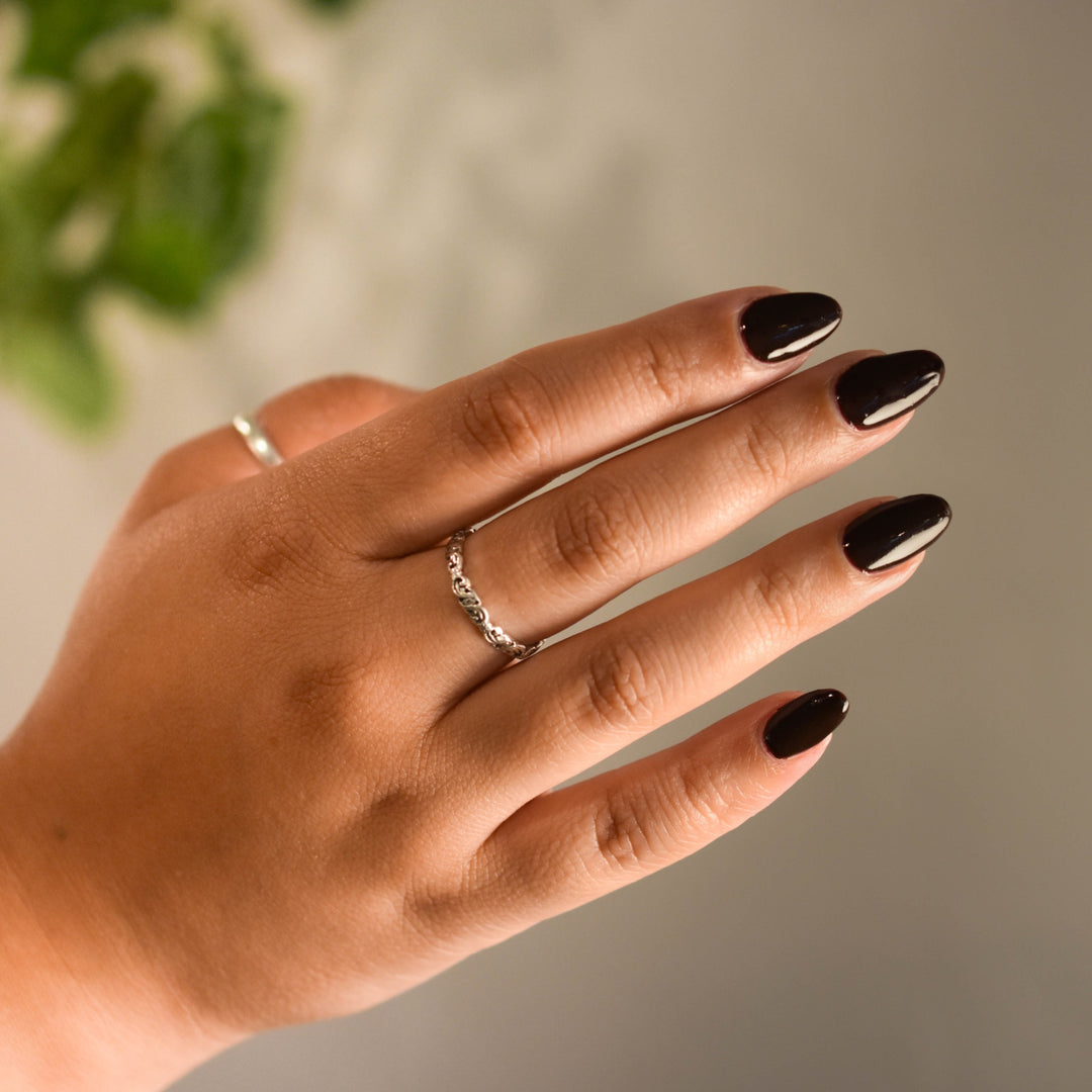 A close up of a medium toned hand wearing Talk Birdie to Me nail polish from Hello Birdie which is a dark oxblood hue. The hand has silver rings on the thumb and middle fingers and floats in front of a white wall with a hanging plant that is out of focus.
