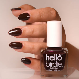 A close up of a hand behind a bottle of Talk Birdie to Me nail polish from Hello Birdie which is a dark oxblood color. The nails are painted with the polish and are glossy. The background is a soft white.