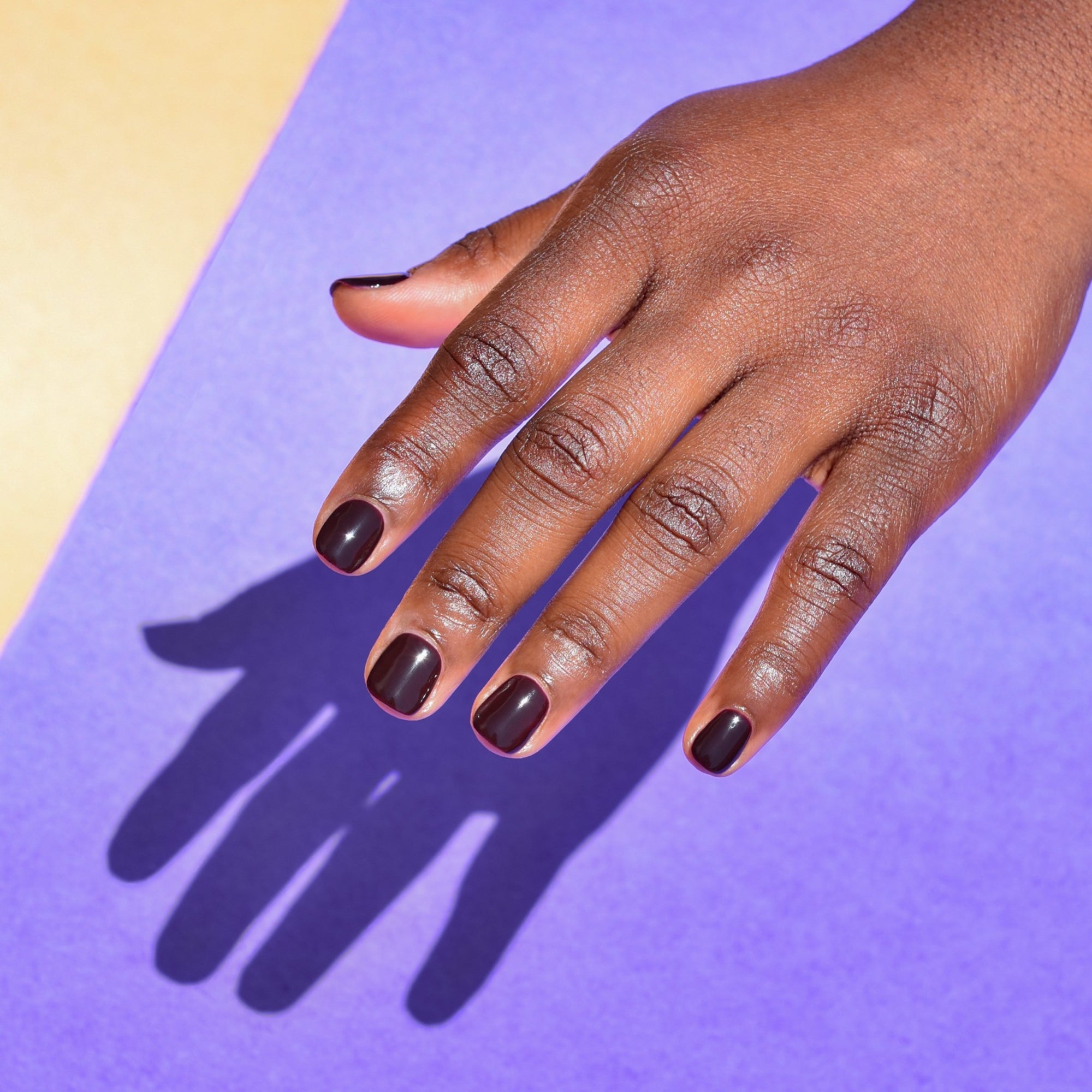 A close up of a brightly lit hand wearing Talk Birdie to Me nail polish from Hello Birdie which is a dark raisin purple hue. The hand casts a sharp shadow against a purple and cream paper background.