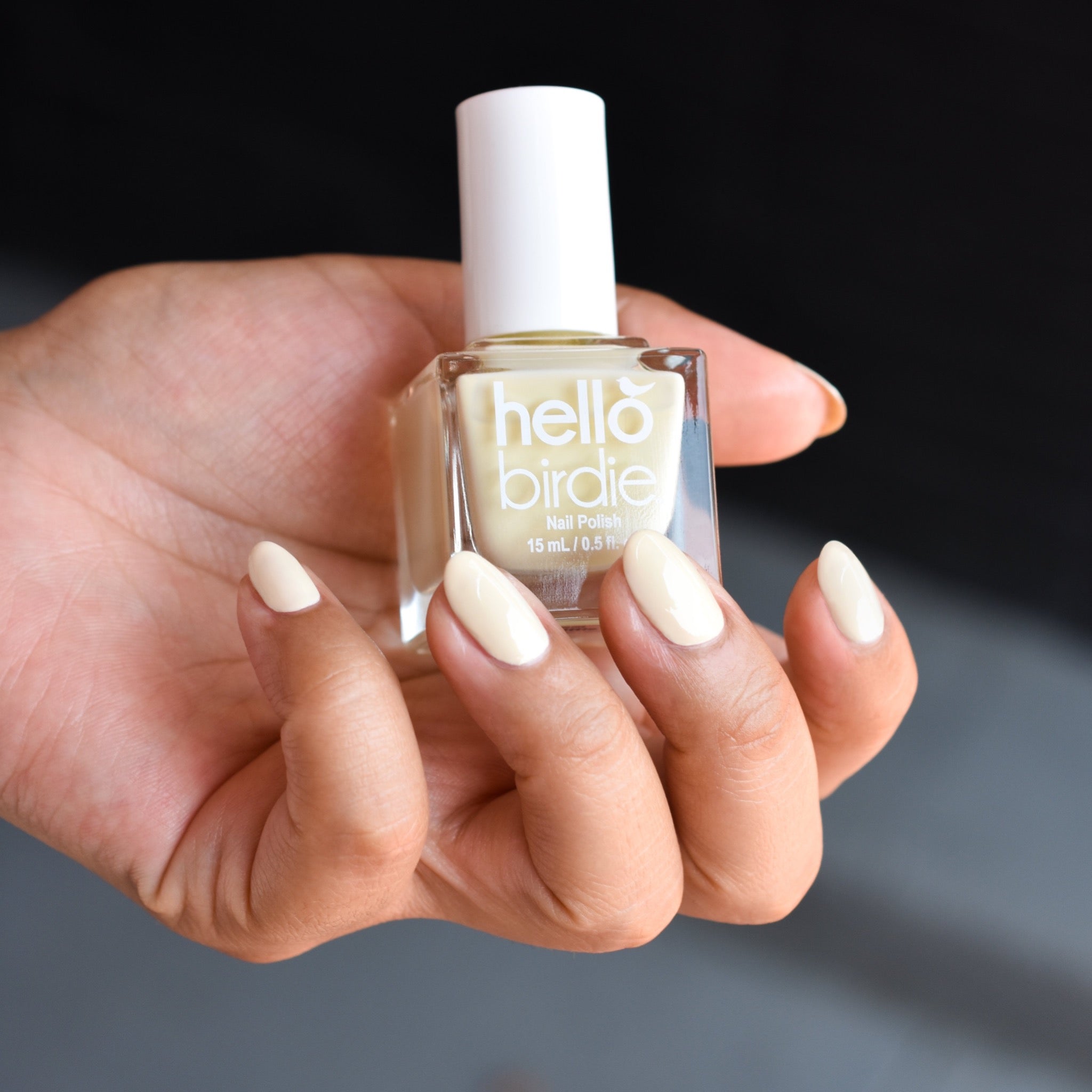 A close up of a mid toned hand comes from the left hand side wearing Three Little Birdies nail polish from Hello Birdie which is a light pastel yellow hue. The hand is clutching a bottle of the nail polish, which has a cube shape and white cap and white text. The background is an out of focus gray and black.