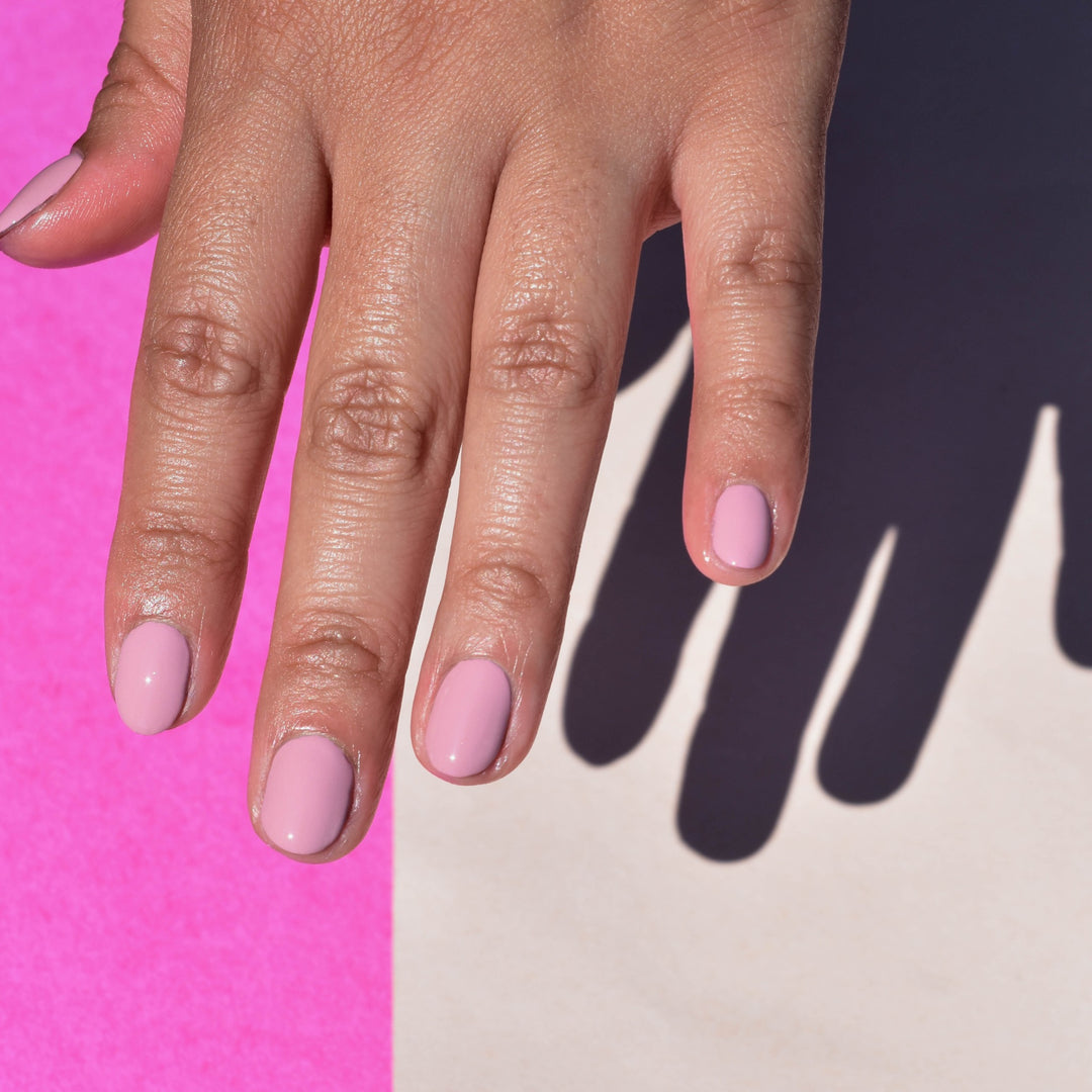A close up of a mid toned hand wearing Tippi nail polish from Hello Birdie which is a rose mauve hue. A hard light illuminates the hand against a two toned bright pink and cream background.