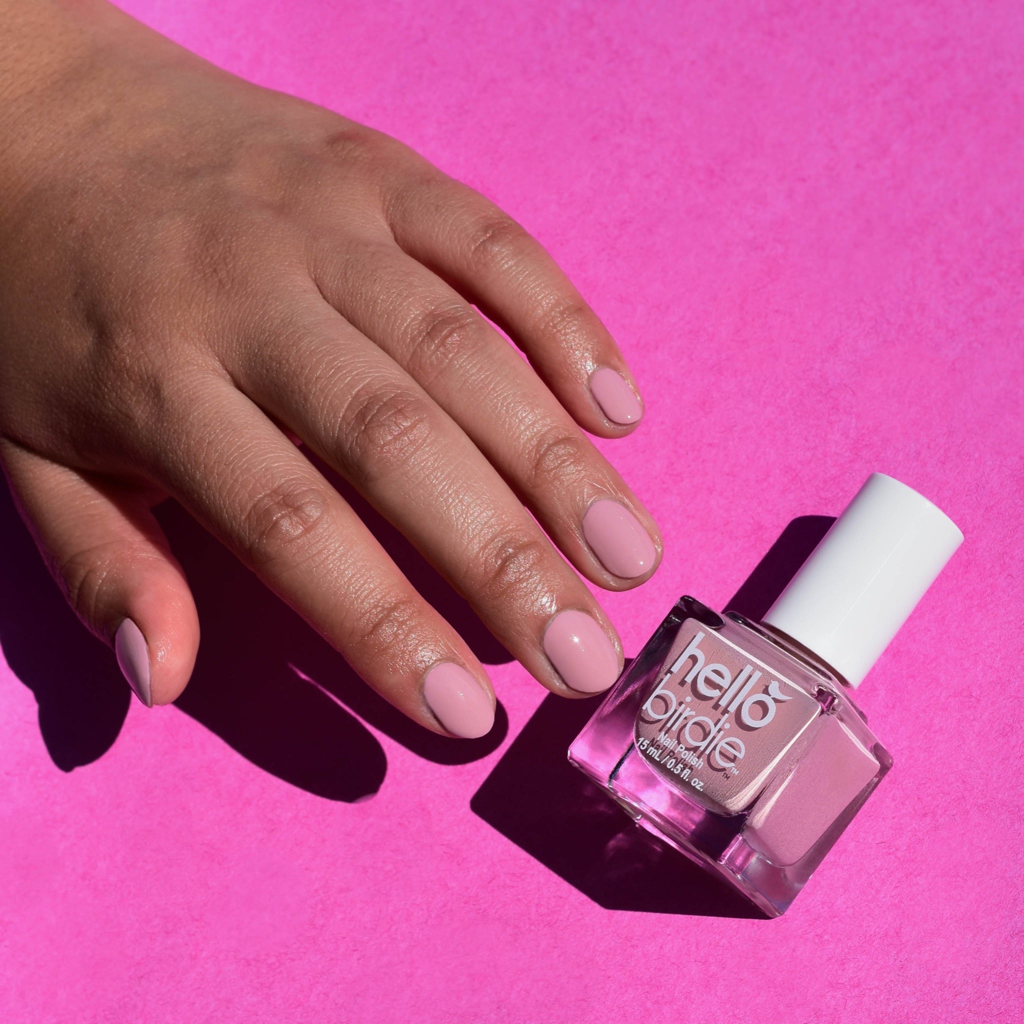 A model's mid toned hand barely touching a bottle of Tippi nail polish from Hello Birdie which is a rosy mauve hue. They are illuminated with a hard light against a bright pink paper background.