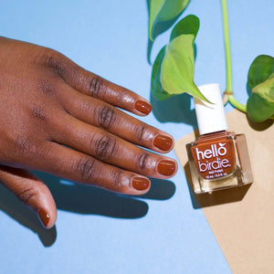 A brightly lit hand hovers next to a bottle of Toucan Play This Game nail polish from Hello Birdie which is a rusty brown hue. The bottle is cube shaped with white cap and text. The fingernails are painted with the same polish and a vine with three leaves comes in from the top while the background is flat sky blue and tan paper.