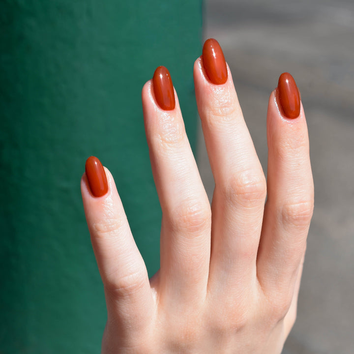 A close up of a light toned hand with Toucan Play This Game nail polish from Hello Birdie in a brownish orange hue. The background is out of focus with a green wall and grey ground.
