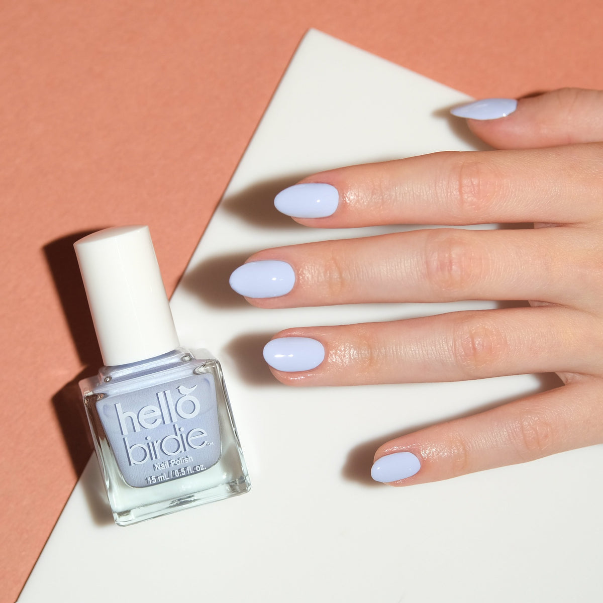 A close up of a light toned hand wearing Tweet Dreams nail polish from Hello Birdie which is a pale blue color. A bottle of the same polish is to the left of the hand and is cube shaped with a white cap and text. The hand is brightly lit in front of a cream and tan offset background.
