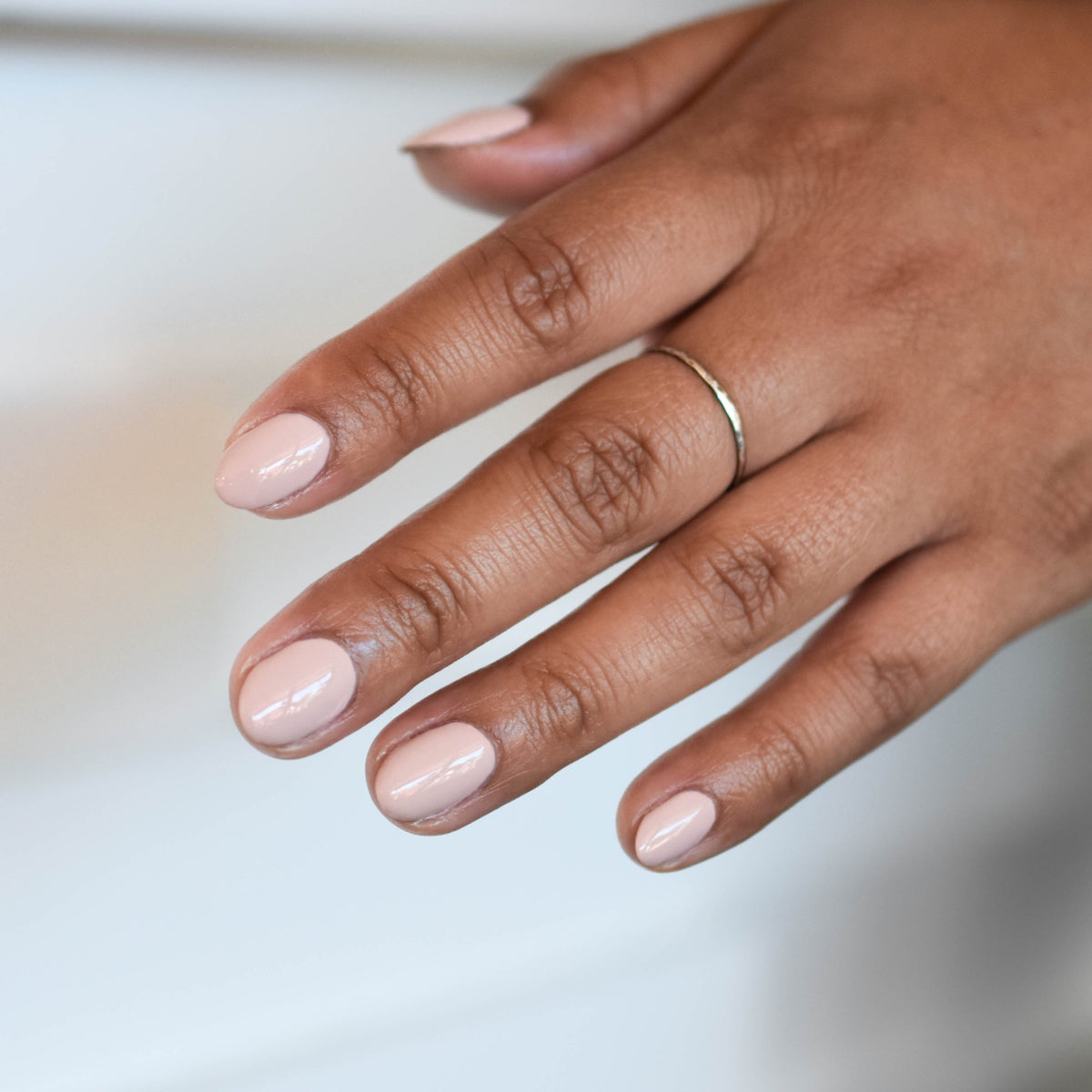 A Close up of a mid toned hand comes run from the upper right hand corner in front of a soft focus white background with soft shadows. The hand has a silver ring on one finger and is wearing Tweet Yo Self nail polish from Hello Birdie which is a beige neutral hue.