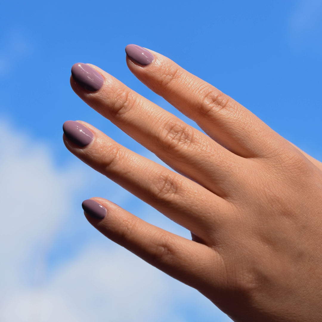 A Close up of a medium toned hand against a blue sky with clouds. The fingernails are painted with Unpheasant nail polish from Hello Birdie in a taupe purple.