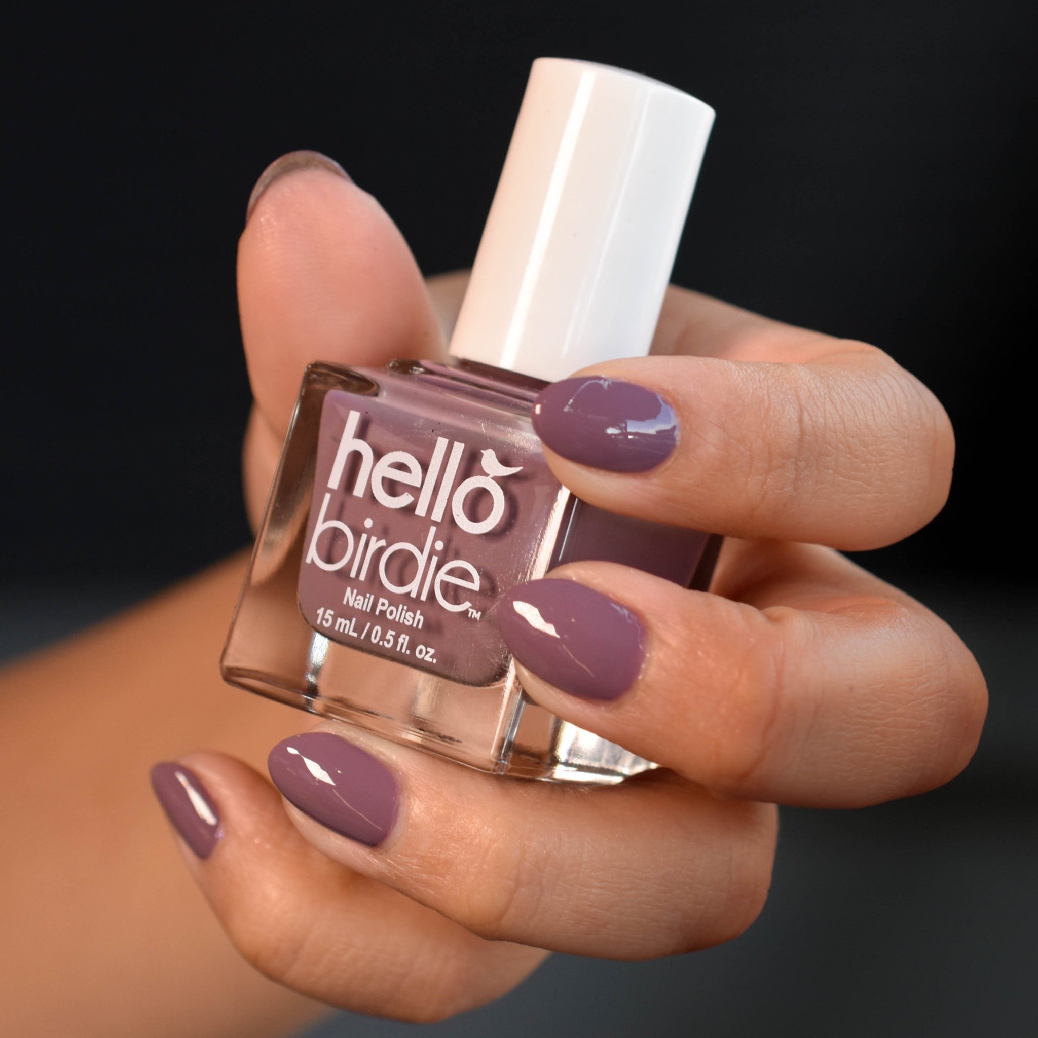 A close up of a medium toned hand clutching a bottle of Unpheasant  nail polish from Hello Birdie in a taupe purple. The nails are painted with the polish and are glossy against a soft black background.