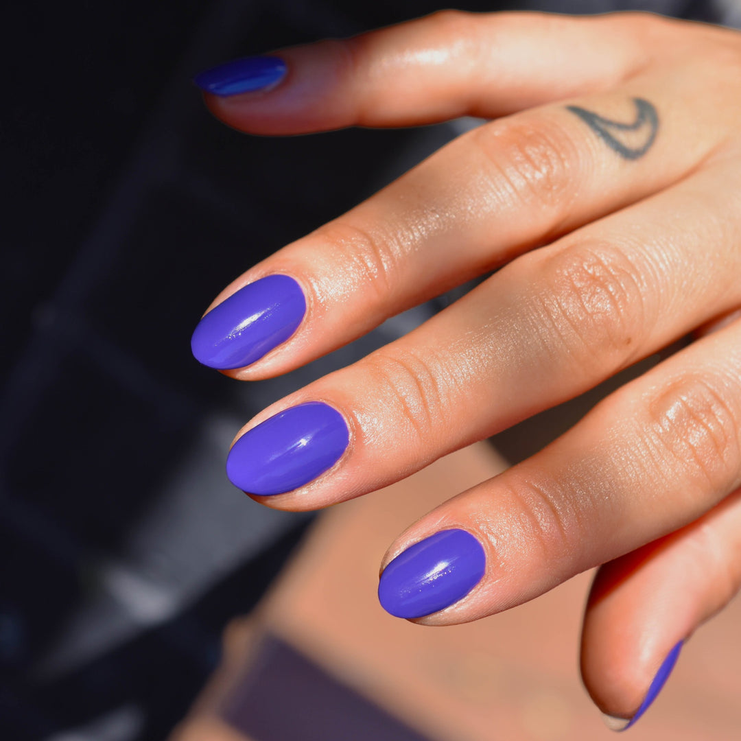 A close-up of a mid toned hand comes in from the upper right hand corner, with daylight shining on the hand but shadows fall across the fingers. There is a moon tattoo on the ring finger and the nails are painted with When Doves Fly nail polish from Hello Birdie in a royal purple hue. An out of focus background displays a black brick wall and tan ground on a tilt.