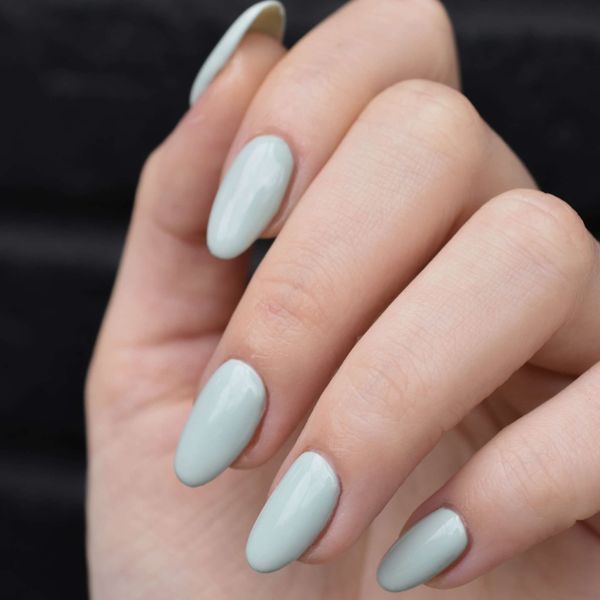A close up of a light toned hand wearing Wingin' It nail polish from Hello Birdie which is a powdery sage green color. The background is an out of focus black brick.