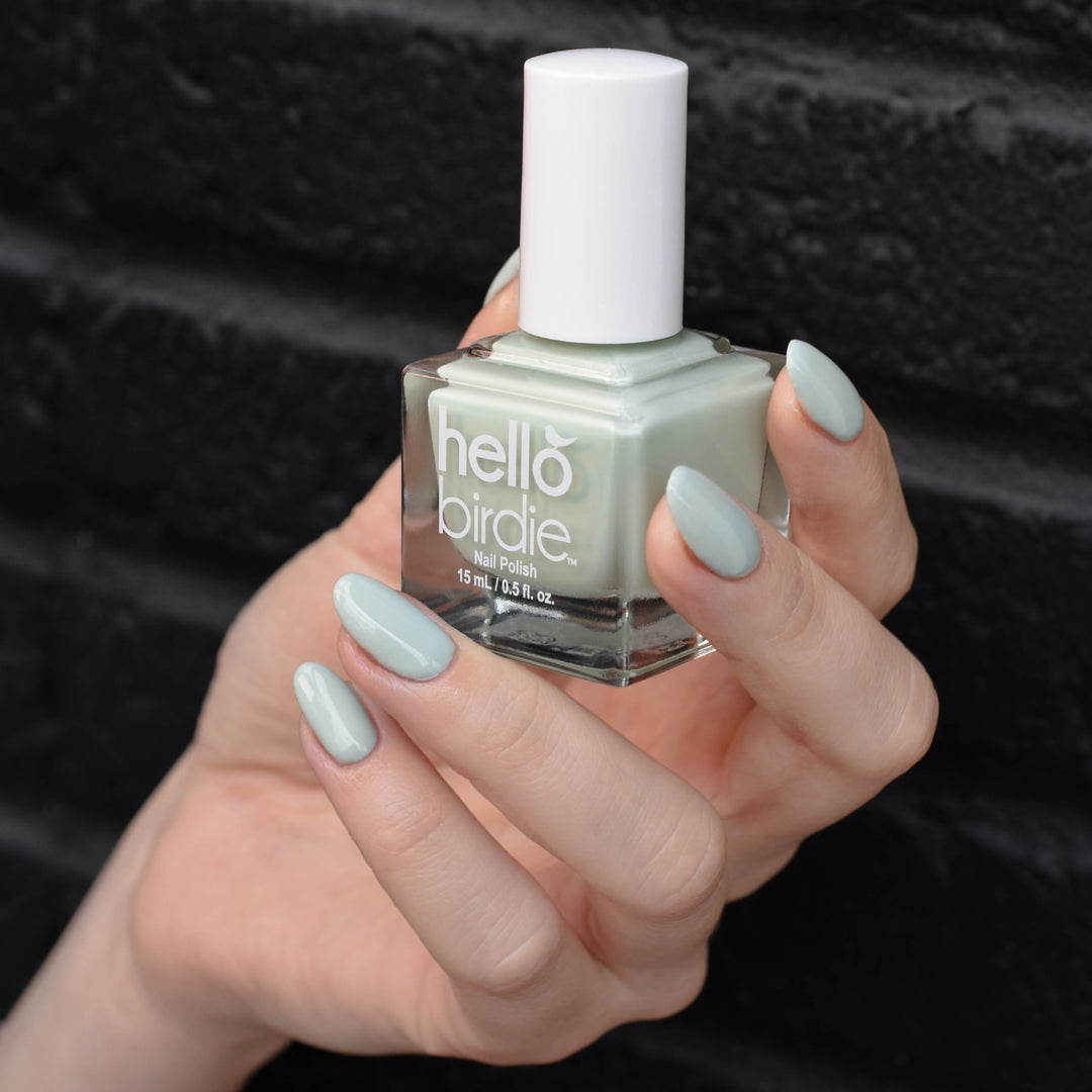 A close up of a light toned hand holding a bottle of Wingin' It nail polish from Hello Birdie in a ;light powdery sage green color. the hand is in front of a black brick background