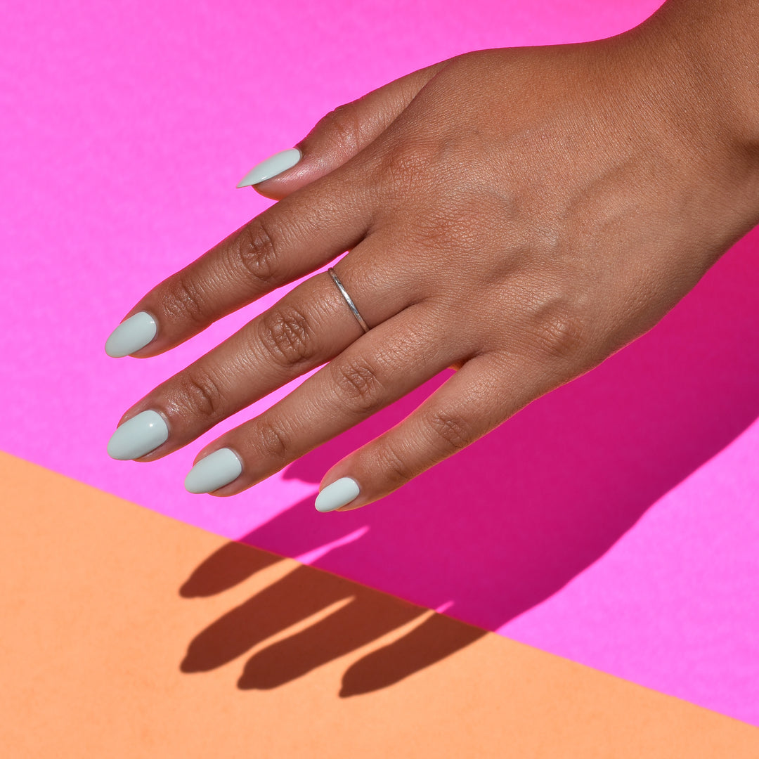 A close up of a hand against a bright pink and tan paper background. The hand is brightly lit and casting a sharp shadow. Wingin' It nail polish from Hello Birdie is painted oil the nails in a light sage hue and the middle finger has a thin silver ring.